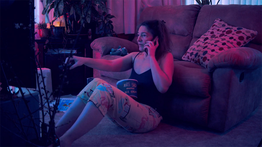 Zuzana is holding phone in one hand and a remote in the other, while she sits disheveled in her pajamas in front of a TV screen. In her lap is a bucket with blue cover and text Anti-sad icecream.